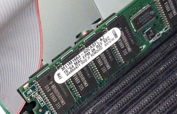 A closeup photo of a RAM DIMM, showing a sticker that reads M11M1664-60CASFL:&Z 16*64 MAC FPM 4K REF SEC 128 MB WARRANTY VOID IF REMOVED 30515732. Four empt sockets are visible in front of it and there is a gray ribbon cable directly behind it.