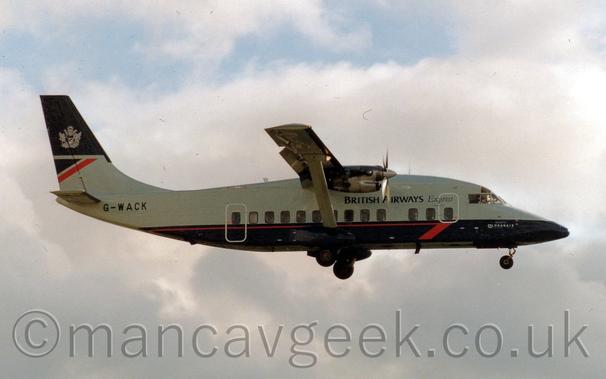 Side view of a boxey, high-winged, twin propellor-engined airliner flying from left to right.
The plane is flying at a low altitude with the undercarriage lowered and flaps extended from the rear of the wings, suggesting it is just about to land.
The plane has a grey fuselage with a dark blue belly, and a red stripe running along the blue part of the body.
There are black "British Airways Express" titles on the upper forward fuselage, the last word being in a thinner, italic font.
The registration "G-WACK" is on the upper rear fuselage, under the tail, also in black.
The top of the tail is dark blue, the rest grey, with a dark blue and red triangle in the middle rear.
Cloudy grey skies with streaks of pale blue peeking through fill the rest of the frame.
