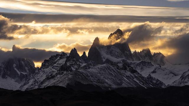 The jagged peaks of Fitz Roy and Cerro Torre rise dramatically above the Patagonian landscape, partially shrouded in swirling clouds and illuminated by golden light. Captured from a viewpoint near El Chaltén in Los Glaciares National Park, Argentina.