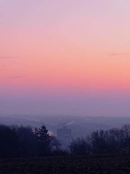 A landscape photo with the upper 2/3 of the  frame being filled by colours of the sunrise. On the bottom third is the outlook on to a landscape from a higher plateau. However, the sight is limited by some fog. In the lower center of the frame a row of trees and bushes make room for a larger apartment building with smoke coming out on top.