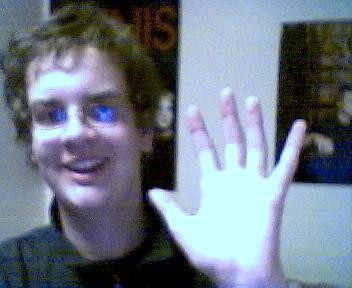 A very low resolution photo of someone with glasses and slightly curly hair waving at a camera, sat in a room with poster on the wall behind them.