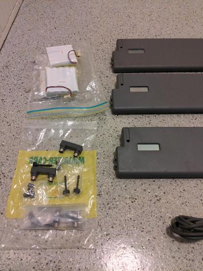 A closeup of four batteries with pigtails (they appear to be 4 AA cells wrapped in white), the left side of three of the dark grey units, showing mounts on the sides and the tiny blank displays. There are also some U-shaped mini-DIN connectors and thumb screws in a ziploc bag.