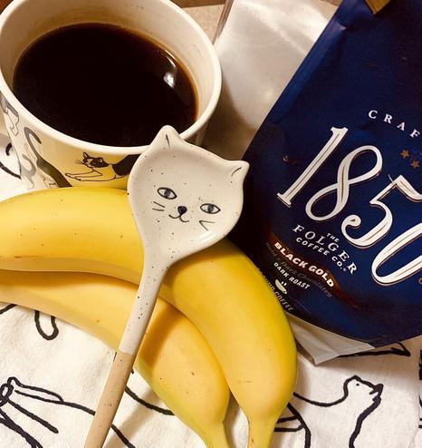 A tea towel with the black outlines of cats is topped with a breakfast spread. A spoon shaped like a cats head, donning a polite smile rest on top of some bananas. A cup of hot coffee is off to the right, and the bag of coffee, 1850 Folgers is on the left.