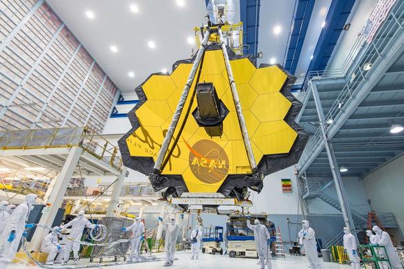 A large, yellow hexagonal satellite mirror is being prepared in a clean room. Several technicians in white protective suits are working around it, while the facility features high ceilings and industrial equipment.