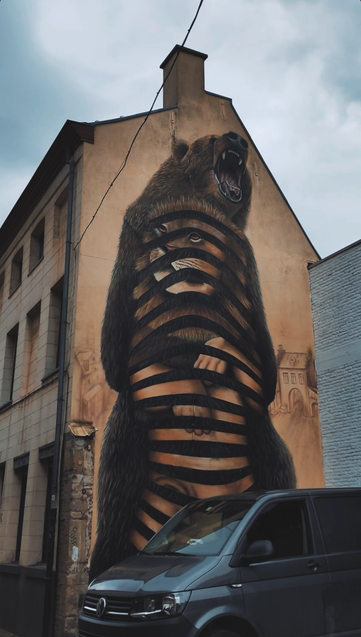 A graffiti in muted colours on the side of a three story building. It is showing a naked human body with a lions head, partially morphing into its shape, nestled inside the body of a standing, roaring bear. Stripes of the bears belly fur is missing, so you can see the humanoid shape within. It has a feral vibe.
