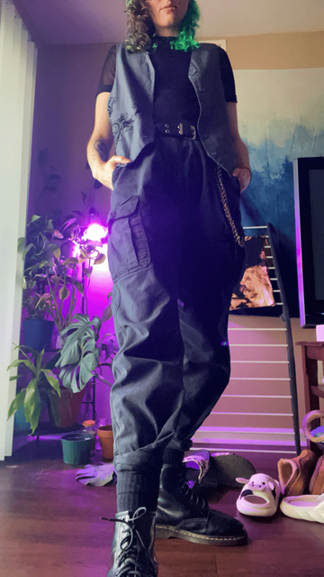 OP in apartment lit up with purple light wearing black Doc Marten boots, grey cargo pants, a black grommet belt, black fishnet shirt, and a grey pinstripe vest with a black floral embellishment on the left pocket.