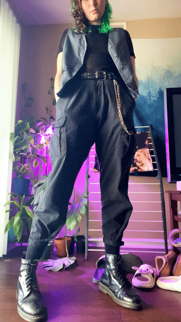 OP in apartment lit up with purple light wearing black Doc Marten boots, grey cargo pants, a black grommet belt, black fishnet shirt, and a grey pinstripe vest with a black floral embellishment on the left pocket.