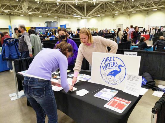 Two volunteers with the Three Waters Pipeline Resistance Team stand behind a table with signs and handouts, talking with an event attendee who is signing a postcard. The setting is a large indoor convention hall at the Minneapolis Convention Center.