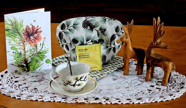 Tea tableau in white, green, and tan with a pinecone motif on linen, cardstock, &yardstick, some carved wooden deer. There is also a crocheted lace doily and a quilted pad. The tea is Sleigh Ride fruit infusion from. David's Teas of Canada.