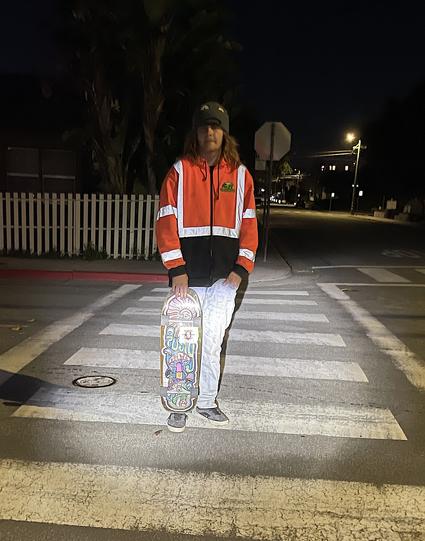 A man with shoulder-length brown hair pauses in a crosswalk on a dark residential street in the city of Monterey. He is looking at the photographer. In his right hand he holds his skateboard, the bottom of which is resting on his right foot. He is dressed in tennis shoes, light blue jeans, a dark-colored brimmed cap,  and a bright orange jacket with high-visibility strips.