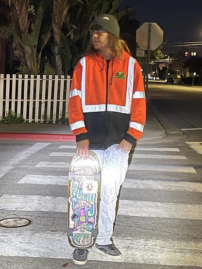 A man with shoulder-length brown hair pauses in a crosswalk on a dark residential street in the city of Monterey. He is looking off to his right. In his right hand he holds his skateboard, the bottom of which is resting on his right foot. He is dressed in tennis shoes, light blue jeans, a dark-colored brimmed cap,  and a bright orange jacket with high-visibility strips.