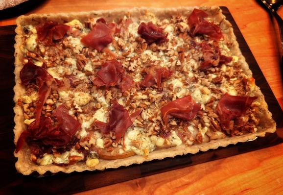 Cooked pear tart topped with prosciutto on cutting board.