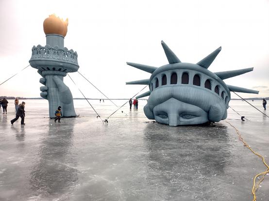 A sculpture in the shape of the top Statue of Liberty on a frozen lake, giving the appearance of the statue being mostly buried in ice.