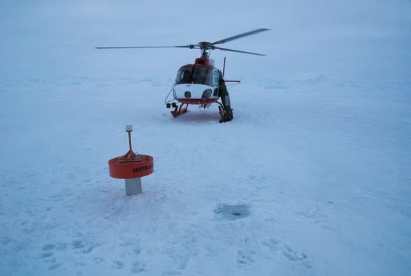 An orange and silver metocean (drift and temperature) buoy stuck into sea ice in the foreground. A red and white helicopter is behind it (Eurocopter AS-350 squirrel), with a person at the rear passenger door. Sea ice extends to the horizon against a cloudy sky