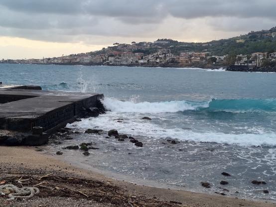 Il mare agitato dal vento alimenta  onde continue che raggiungono la riva. In lontananza sotto un cielo grigio, si affacciano case costruite sulla costa