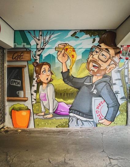 Streetartwall. A very funny and well-painted comic mural showing a man and his girlfriend in different stages of life was sprayed/painted on a wall in Berlin. The bearded man with short brown hair and glasses stands there in a gray baggy sweater and holds a pizza box under his arm. With his other hand, he holds up a slice of pizza from which the cheese is dripping and which he is clearly looking forward to. In the background, a horrified young woman with a ponytail hairstyle in a pink and gray sports outfit can be seen doing her yoga exercises on a meadow. She looks jealously at her boyfriend, who is eating the delicious slice of pizza. Next to it is a café kiosk with a cup of coffee on the counter and a flower pot.
Info: The artist on his magnificent work of art: "Tried to paint a typical situation between the love of my life and me. She's doing her fav thing while I'm eating delicious stuff and she's becoming jealous about it."