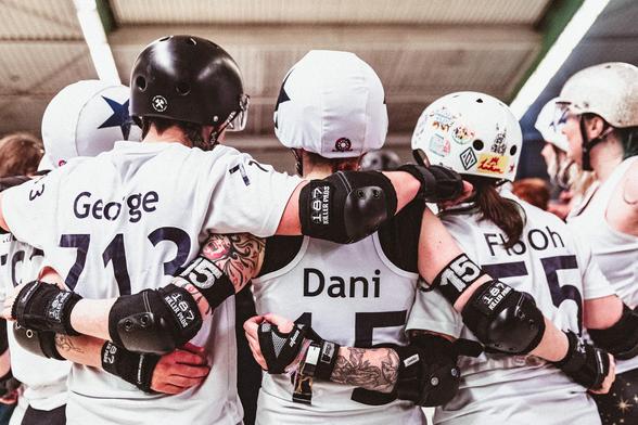 Subdued colour picture of Roller Derby playing people. Three players standing back to camera with their arms wrapped around each other, listening to a person in front of them (not visible)