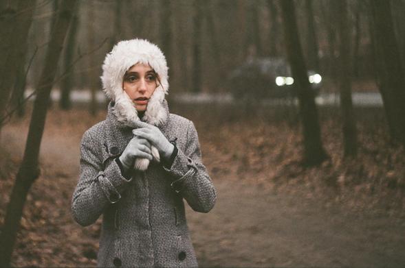 The image shows a person wearing a winter coat with a hood that has a furry lining. They are also wearing gloves and appear to be in a forest or wooded area. There is a vehicle with headlights on in the background, suggesting a road nearby. The scene looks cold and the setting is likely outdoors during winter or late autumn.