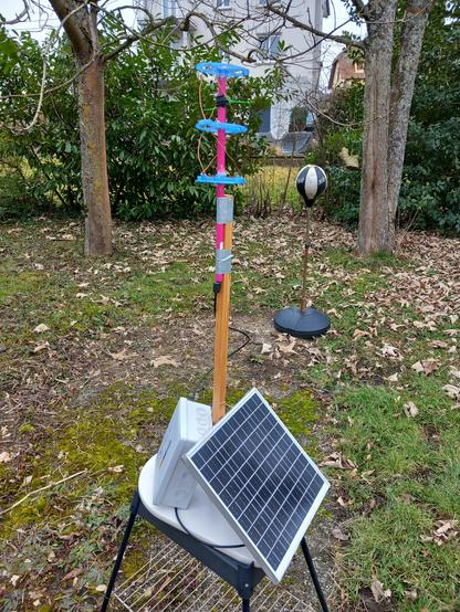 Une station-sol constituée d'un panneau solaire de petite taille sur la droite, d'un boîtier électrique gris IP55 sur la gauche, et d'un poteau en bois supportant une antenne quadrifilaire DIY. Le support de l'antenne est rose fluo alors que les flasques horizontaux sont bleus. L'ensemble est dans un jardin vert avec de nombreuses feuilles sèches au sol.