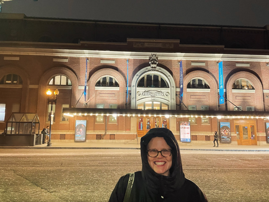Woman standing in front of symphony hall wearing a winter coat with the hood up