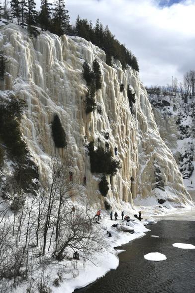 A cliff covered in ice, with conifers above and cedar trees growing on its side. Four ice climbers scale the wall. Below, others ready for their own climb and steady the safety ropes for the climbers, next to the river which remains open with some patches of ice along its banks.