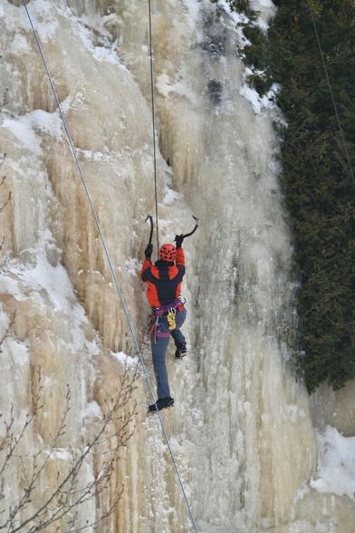 An ice climber scales a wall of yellowed ice next to a cedar tree that grows from the cliffside. Their right foot is raised to their knee, with crampons dug into the ice. Hanging from the ice pick in their left hand, they reach high with their right arm, swinging a second ice pick. They’re also equipped with a helmet, harness, carabiners, and extra rope.