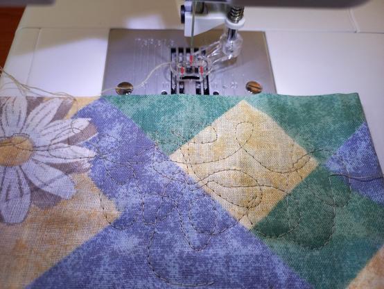 A piece of multicolored scrap fabric, laying on the sewing machine, just in front of the presser foot. There are uneven squiggly lines sewed with a straight stitch. The lines are done with free motion sewing which causes stitch length to be varied as I move the fabric my hand while the needle does its work with forming stitches.