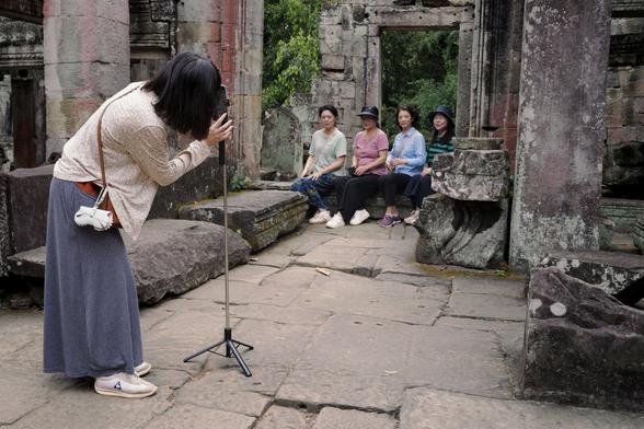 Eine Frau fotografiert mit einem Smartphone auf einem Stativ eine Gruppe von vier Frauen, die auf einer Steinbank vor antiken Ruinen sitzen. Die Szene spielt sich in einer grünen, historischen Umgebung ab. (Angkor Wat, Preah Khan)