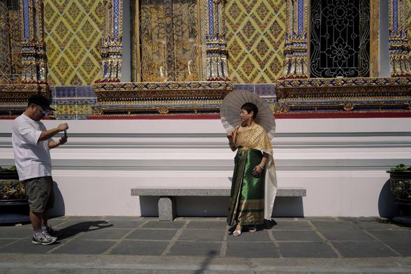 Ein Mann in Freizeitkleidung fotografiert eine Frau, die traditionelle Kleidung und einen Sonnenschirm trägt. Sie posiert vor einer reich verzierten Wand mit bunten Mustern und goldenen Details. (Bangkok, Wat Arun)