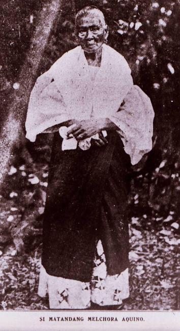 A monochromatic photograph of an elderly Aquino, standing against a blurred backdrop of trees. She wears a white garment with wide sleeves & a dark lower garment. Her face shows a gentle expression, she holds a small item in front, with shadows ^ highlights creating depth in the composition. The image has a grainy texture typical of historical photographs.