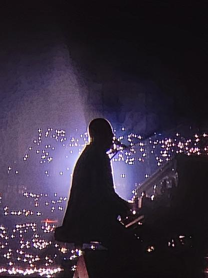 Silhouette of Billy Joel playing the piano backlit by the audience holding lights at a live show.