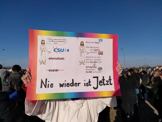 Ein Mann in weißer Tunika trägt ein Schild.

Links Jesus (in weißer Tunika): Sorry, aber das muss ein Misverständnis sein. CSU Logo, aber C für Christlich und S für Sozial sind durchgestrichen.

Rechts nochmal Jesus: Recht auf Asyl, Alle Menschen sind gleich wertvoll, Gerecht ist nicht was mir, sondern was ALLEN nützt, Schöpfung bewahren, KLIMA schützen.

Unten groß: Nie wieder ist Jetzt