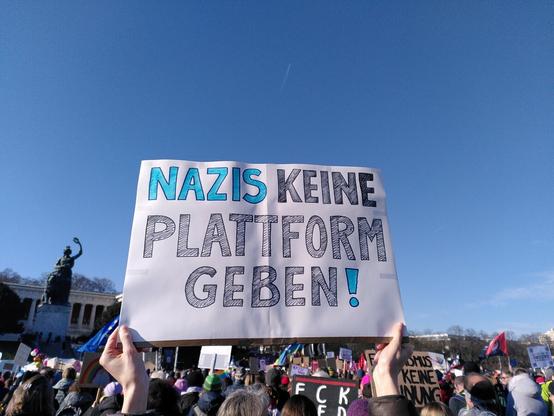 Schild: NAZIS KEINE PLATTFORM GEBEN. Hinter dem Schild sieht man viele Köpfe und die Bavaria