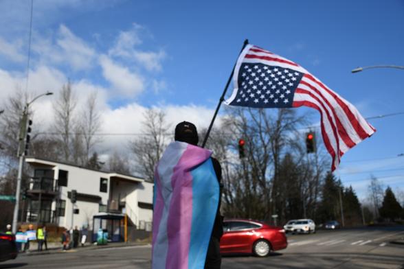 A protestor wearing a trans pride flag. They are waving the American flag, which has been flipped upside down, in the direction of commuters.