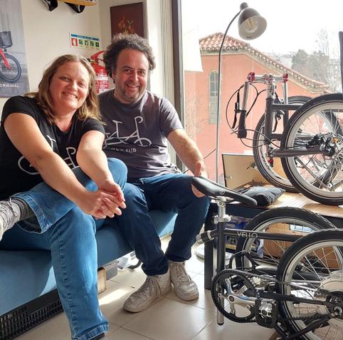 Um casal, homem cabelo caracóis, mulher cabelo loiro, ambos com t-shirts preta com bicicleta, fundadores da marca de bicicletas VELLO. Sentados num sofá azul dentro da loja de bicicletas junto a duas bicicletas dobráveis VELLO