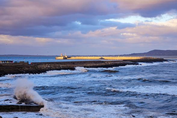 a stone pier stretching g
into the distance. the scene is cloudy but the far end of the pier is lit by sunshine. waves break on a smaller pier in the foreground