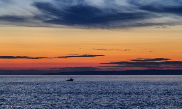 Looking across a bay at sunset, with dark water in the foreground and an orange and gold sunset with dark wispy clouds above and a few dark streaks of clouds on the horizon. There are low hills on the far shore of the bay. A lone fishing boat with a light is just about in the centre of the image, where the water meets the opposite shore.