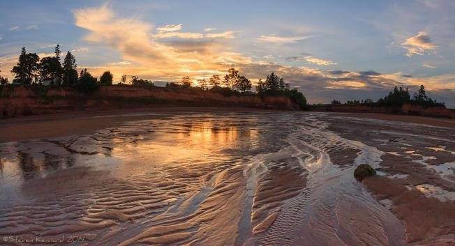 A sunset scene looking from the water's edge up an expanse of dark red undulating sand with ripples and rivulets coming down from the shore. The sunset is reflected in the surface water and almost mirrors the sky. The dark skyline is low cliffs with trees at the top. The sky, between the wisps of cloud is blue.