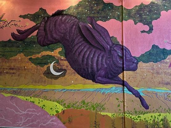 A purple jack rabbit leaps over the crescent moon, the sky is filled with pink clouds and stars, cliffs and canyon lands below.