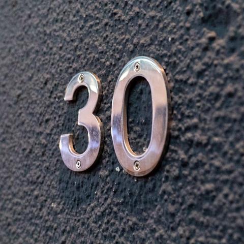 Number 30 in metal, screwed into a grey textured outer wall, at a slight perspective angle with right, closer to left