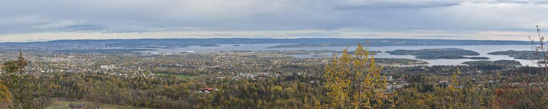 A panoramic photo looking over Bærum and the Oslofjord from the top of a hill. The sky is cloudy.