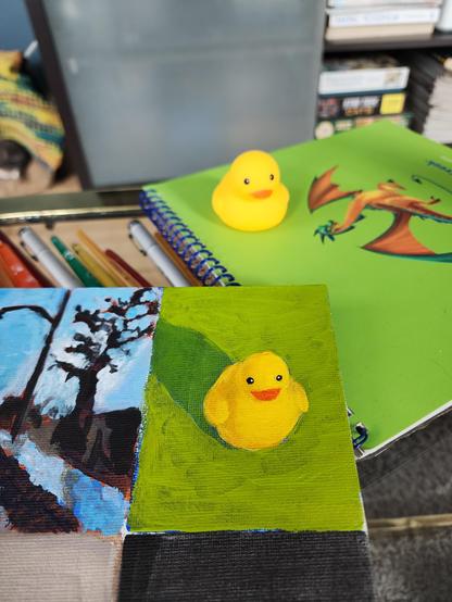 An acrylic painting of a yellow rubber ducky on a bright green background. On the table behind the painting is the actual duck sitting on a bright green notebook