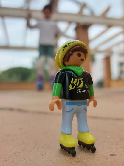 Criança playmobil de patins e gorro amarelos. Camisola preta. Calças azuis claras. Atrás uma criança real de patins