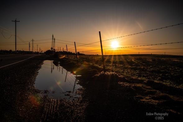 Off to the side of the roadway on the right side of this scene, the sun rises between the strings of barbed wire.  On the left side, a long puddle left over by the recent rains, separate the barbed wire fence and the roadway.