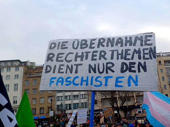 Auf dem Ebertplatz hält jemand ein Schild hoch: "Die Übernahme rechter Themen dienst nur den Faschisten", im Hintergrund Demonstrant*innen mit Fahnen und Schildern.