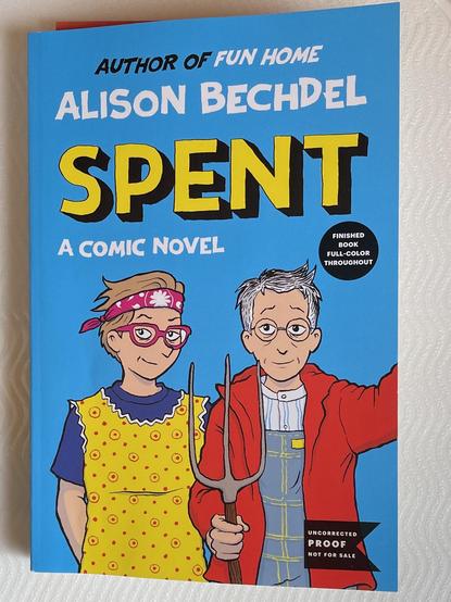 Photo of the book, cover illustration is a bright blue background with cartoon drawing of Alison Bechdel and Holly Rae Taylor in the style of American Gothic, with Alison as the man and Hol as the wife. There are two sticker-looking callouts, the first reads, “finished book full-color throughout” and the second reads “uncorrected proof not for sale”