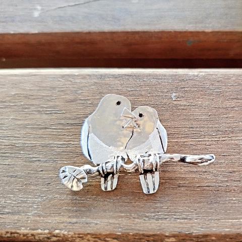 A silver pendant with two loving birds. it has a black cord. it's placed on piece of wood