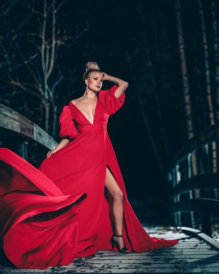 The image depicts a woman standing on a wooden bridge at night. She is wearing a flowing red dress with dramatic sleeves and a high slit. The background is dark, with barren trees visible, suggesting a winter or late autumn setting. She is posing with one hand on her head, and the dress is elegantly spread out, adding a sense of movement and grace to the scene.