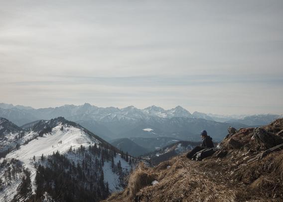 person sitting on a hillside in the left foreground while looking into the far distance with snowy mountain peaks in the distant