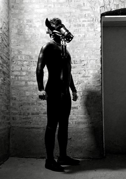 The black-and-white image captures an human pony. The figure is clad in a sleek silicone suit, emphasizing the contours of their body, and wears a leather hood designed to evoke equine features. This hood incorporates elements such as straps, a muzzle, and D-rings, which are common in pony play gear to enhance the visual and sensory experience.

Set against a raw brick backdrop, the composition evokes an industrial yet intimate atmosphere. The interplay of light and shadow highlights the figure's presence while underscoring themes of power and transformation central to pony play.